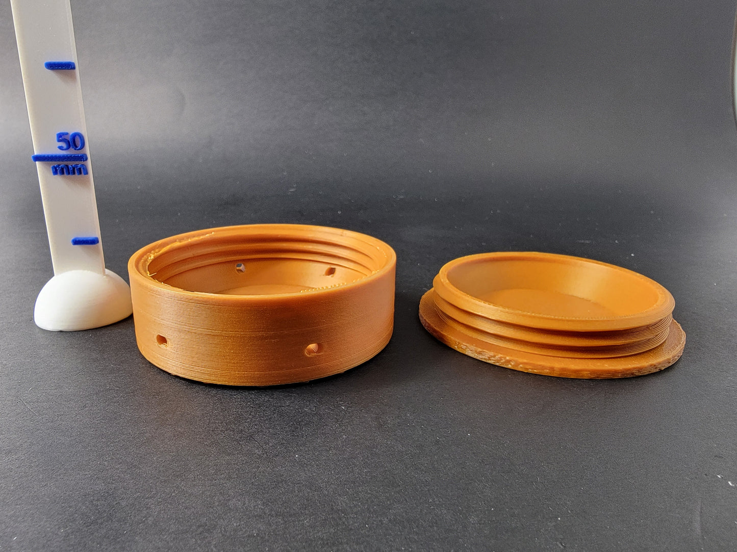 Bronze 3D printed medal hide open and disassembled next to a 50mm scale ruler, showing interior scent chamber